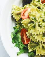 A white bowl filled with bow-tie pasta coated in a vibrant green pesto sauce, garnished with fresh cherry tomato halves and nestled on a bed of mixed greens.