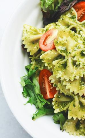 A white bowl filled with bow-tie pasta coated in a vibrant green pesto sauce, garnished with fresh cherry tomato halves and nestled on a bed of mixed greens.